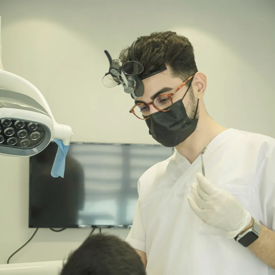 Dentista con lupas y mascarilla atendiendo a un paciente.